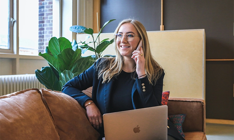 Woman in blue long sleeve shirt using silver macbook and calling on her phone.